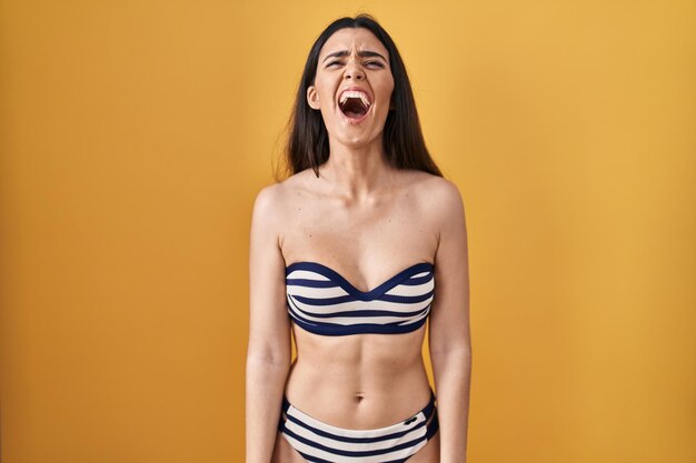 Young brunette woman wearing bikini over yellow background angry and mad screaming frustrated and furious shouting with anger rage and aggressive concept