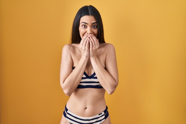 Free photo young brunette woman wearing bikini over yellow background laughing and embarrassed giggle covering mouth with hands, gossip and scandal concept