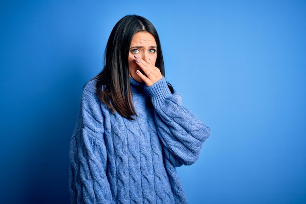 무료 사진 캐주얼 터틀넥 스웨터를 입은 파란 눈을 가진 젊은 브루네트 여성은 코에 손가락을 대고 숨을 참는 냄새나고 역겨운 냄새를 풍깁니다.