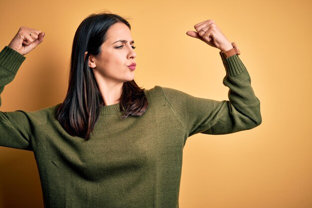 Young brunette woman with blue eyes wearing green casual sweater over yellow background showing arms muscles smiling proud Fitness concept