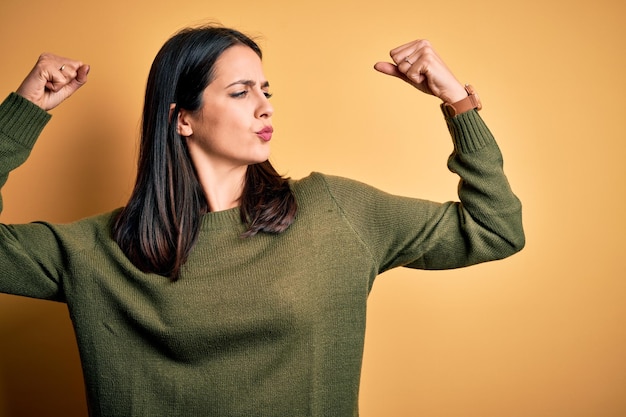 Young brunette woman with blue eyes wearing green casual sweater over yellow background showing arms muscles smiling proud Fitness concept