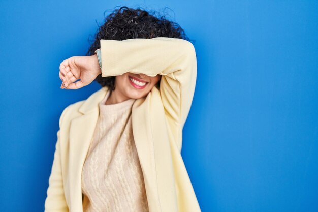 Young brunette woman with curly hair standing over blue background covering eyes with arm smiling cheerful and funny. blind concept.