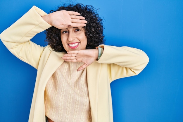 Foto gratuita giovane donna castana con capelli ricci in piedi su sfondo blu sorridente gioco allegro sbirciare un fischio con le mani che mostrano il viso sorpreso ed uscito