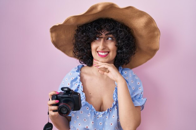 Young brunette woman with curly hair using reflex camera serious face thinking about question with hand on chin, thoughtful about confusing idea