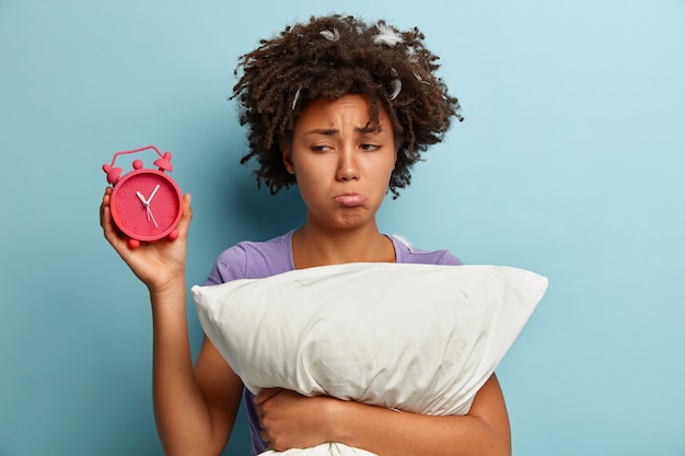 Free photo young brunette woman with feathers in hair holding pillow and alarm clock