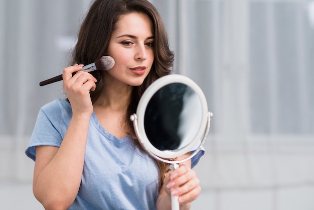 鏡で見ている化粧ブラシを持つ若いブルネットの女性