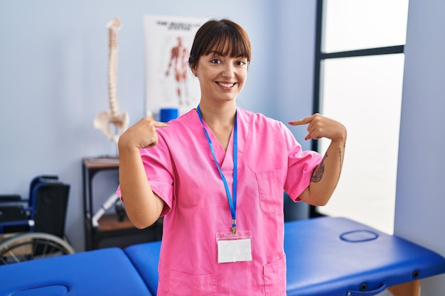 Free photo young brunette woman working at rehabilitation clinic looking confident with smile on face pointing oneself with fingers proud and happy