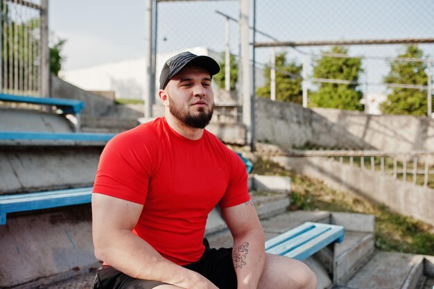 Young brutal bearded muscular man wear on red shirt shorts and cap at stadium