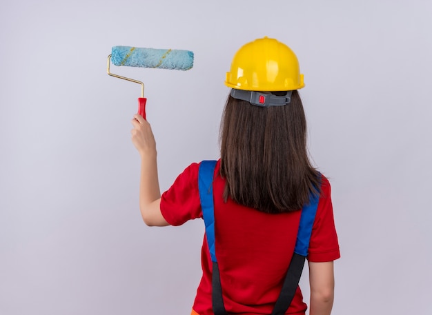 La giovane ragazza del costruttore sta con la parte posteriore alla macchina fotografica e tiene il rullo di vernice su fondo bianco isolato