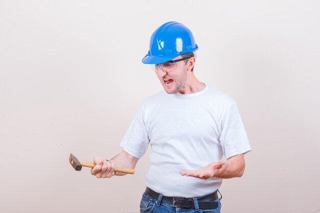 Молодой строитель держит молоток во время крика в футболке, джинсах и шлеме