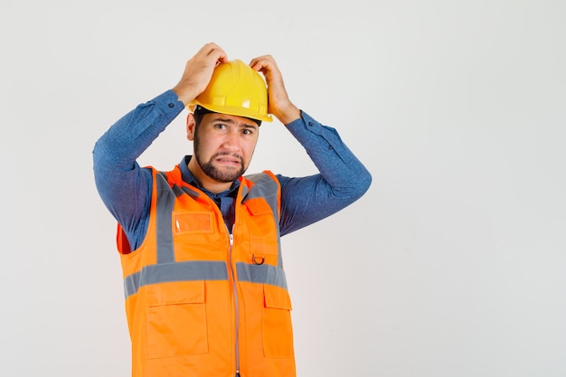 Young builder holding hands on head in shirt, vest, helmet and looking helpless , front view.