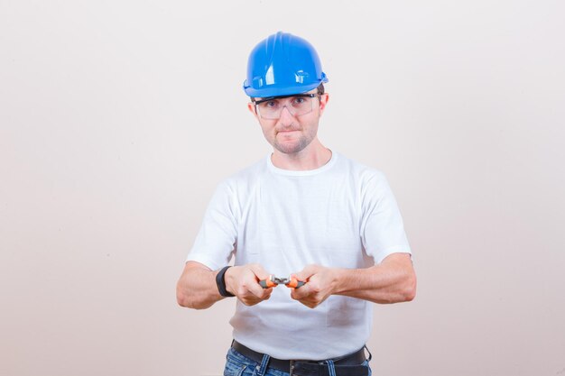 Young builder holding pliers in t-shirt, jeans, helmet and looking confident