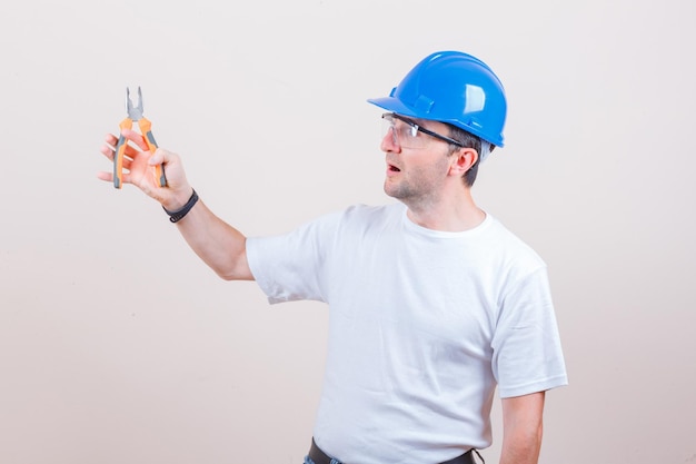 Young builder holding pliers in t-shirt, jeans, helmet and looking surprised