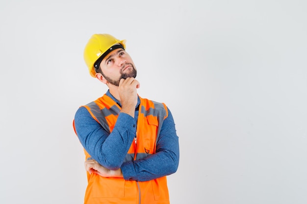 Young builder looking up in shirt, vest, helmet and looking pensive , front view.