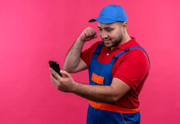 Young builder man in construction uniform and cap going to punch his mobile angry and frustrated 