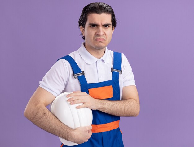 Young builder man in construction uniform holding his safety helmet looking at front sad expression making wry mouth standing over purple wall