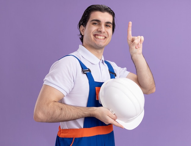 Young builder man in construction uniform holding his safety helmet looking at front with confident expression pointing with index finger up smiling standing over purple wall