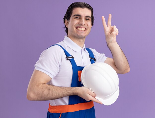 Young builder man in construction uniform holding his safety helmet looking at front with confident smiling showing v-sign standing over purple wall