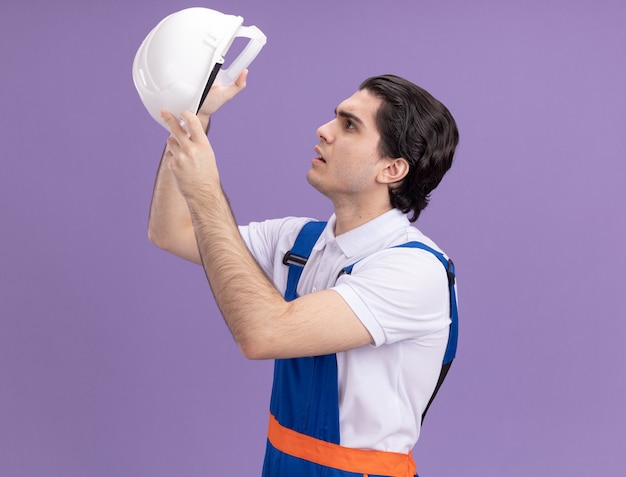 Young builder man in construction uniform holding his safety helmet looking at it intrigued standing over purple wall