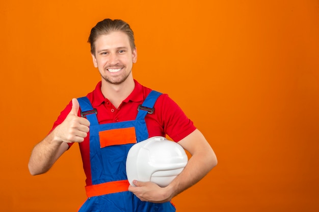 Молодой строитель человек в строительной форме, держа в руке защитный шлем и показывает палец вверх с большой улыбкой на лице, стоя над изолированной оранжевой стене с копией пространства