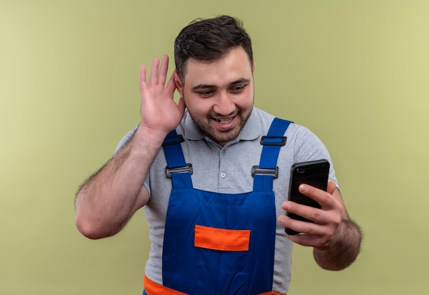 Young builder man in construction uniform holding smartphone holding hand near ear trying to listen  