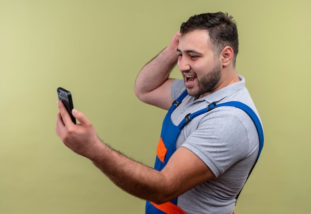 Young builder man in construction uniform holding smartphone looking at screen connecting with someone  