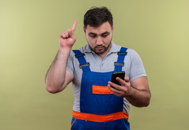 Foto gratuita uomo giovane costruttore in uniforme da costruzione tenendo lo smartphone guardando lo schermo puntare il dito verso l'alto ricordando a se stesso