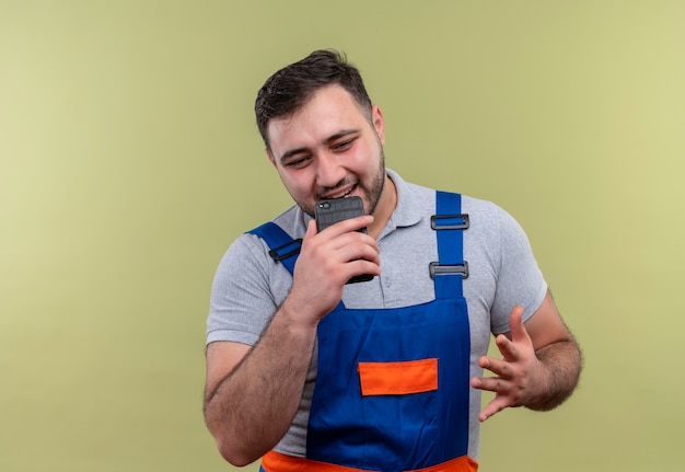 Young builder man in construction uniform holding smartphone sending voice message to someone  