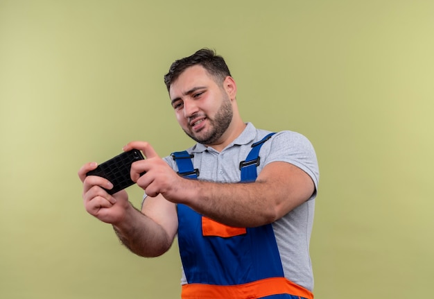 Uomo giovane costruttore in uniforme di costruzione che gioca gioco sul suo smartphone