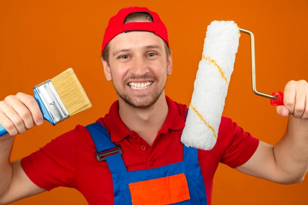 Young builder man in construction uniform and red cap holding paint roller and brush with big smile on face over isolated orange wall