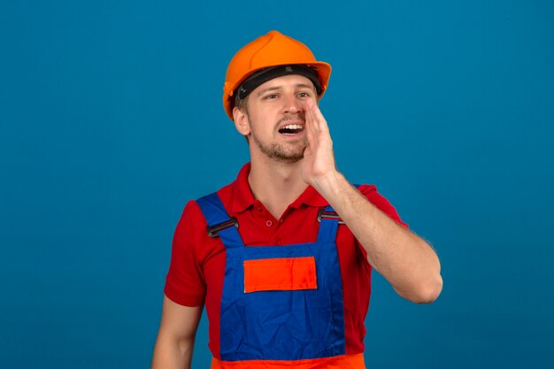 孤立した青い壁の上の口の近くの手で誰かを呼び出す建設ユニフォームと安全ヘルメットの若いビルダー男