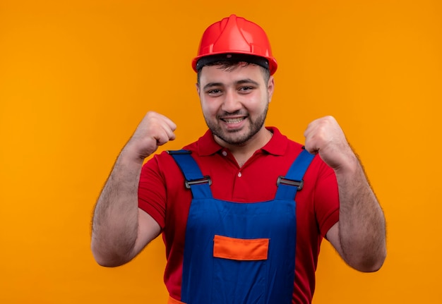 Young builder man in construction uniform and safety helmet clenching fists happy and exited rejoicing his success 