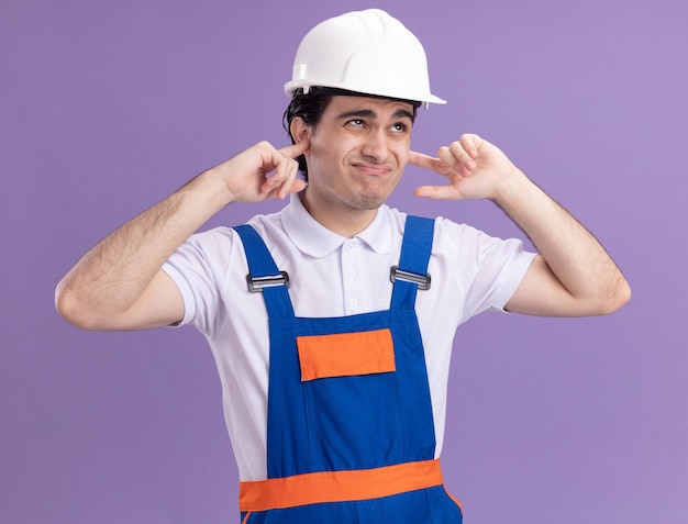 Uomo giovane costruttore in uniforme da costruzione e casco di sicurezza chiudendo le orecchie con le dita con espressione infastidita in piedi sopra la parete viola