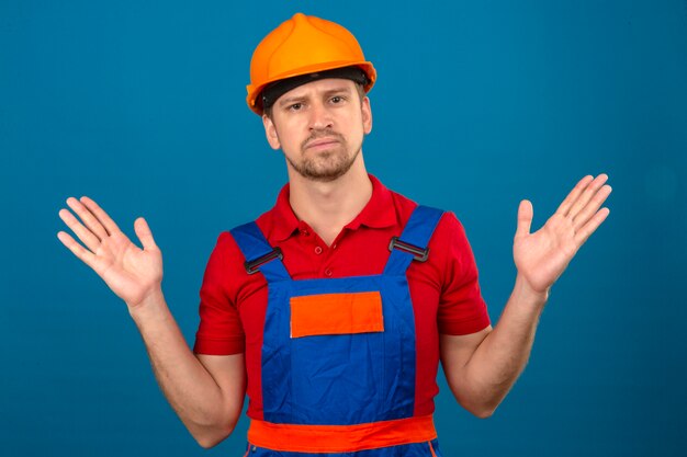 Молодой строитель в строительной форме и защитном шлеме с невежественным и растерянным выражением лица с руками и руками вызывал сомнение в концепции над изолированной синей стеной