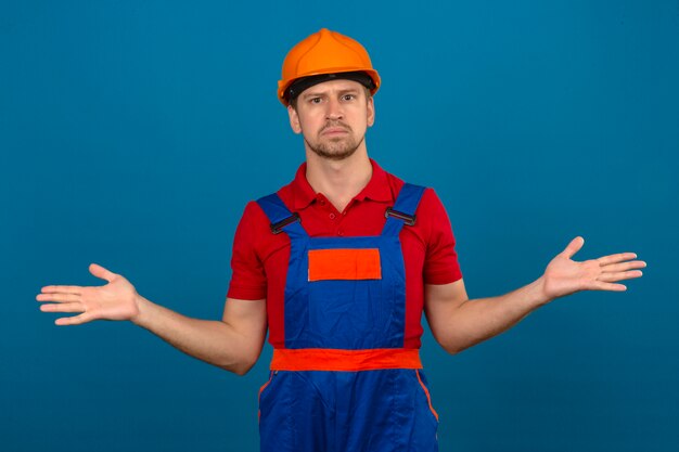 Young builder man in construction uniform and safety helmet clueless and confused expression with arms and hands raised doubt concept over isolated blue wall