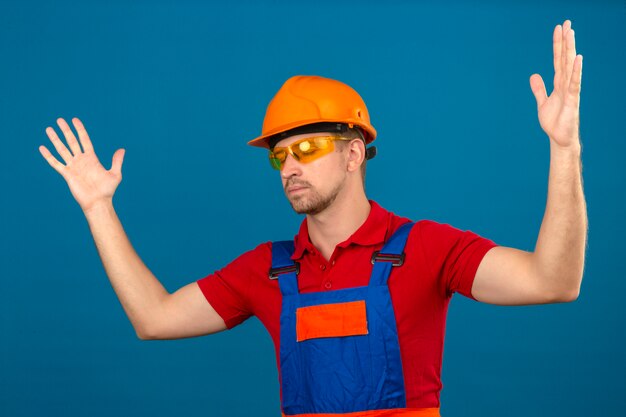 Young builder man in construction uniform and safety helmet gesturing disappointed over isolated blue wall