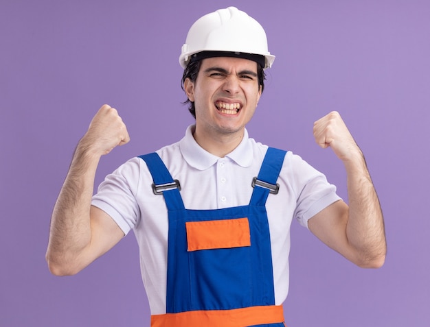 Молодой строитель в строительной форме и защитном шлеме счастлив и взволнован, сжимая кулаки, радуясь своему успеху, стоя над фиолетовой стеной