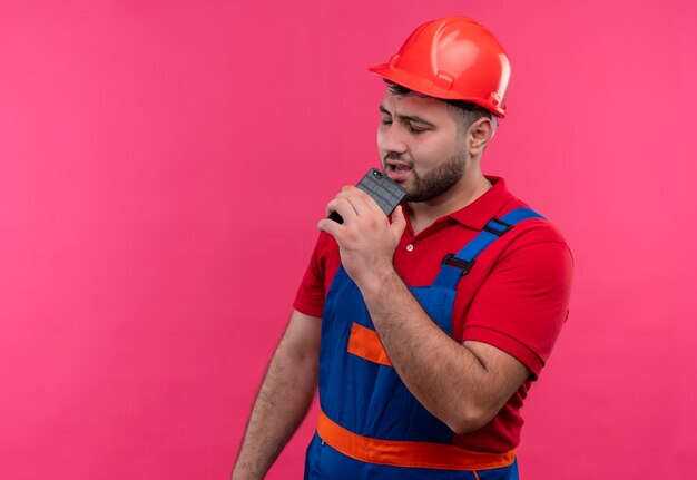 Young builder man in construction uniform and safety helmet hlding smartphone using it as microphone singing 