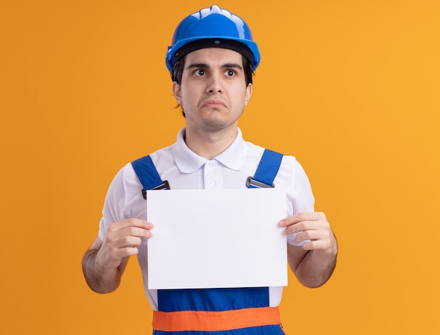 Молодой строитель в строительной форме и защитном шлеме держит пустую страницу, глядя в сторону с грустным выражением лица, стоящего над оранжевой стеной