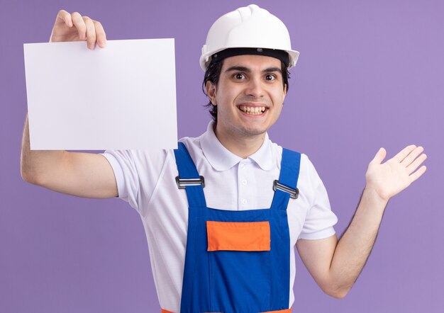 Young builder man in construction uniform and safety helmet holding blank page looking at front with smile on face standing over purple wall