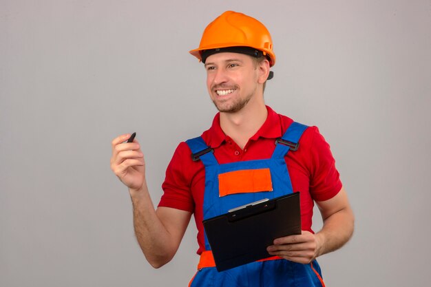 Молодой строитель в строительной форме и защитном шлеме с буфером обмена и ручкой, глядя в сторону с улыбкой на лице над изолированной белой стеной