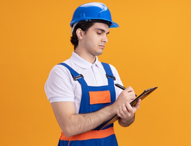 Young builder man in construction uniform and safety helmet holding clipboard writing with pen looking confident standing over orange wall