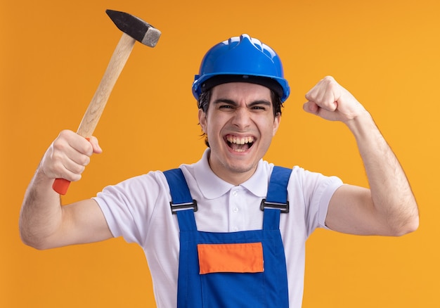 Young builder man in construction uniform and safety helmet holding hammer looking at front happy and excited clenching fist standing over orange wall