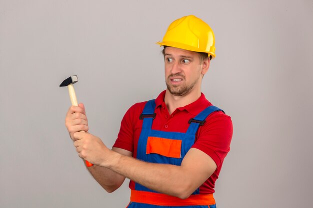 Young builder man in construction uniform and safety helmet holding hammer and looking at it with skeptical face expression over isolated white wall