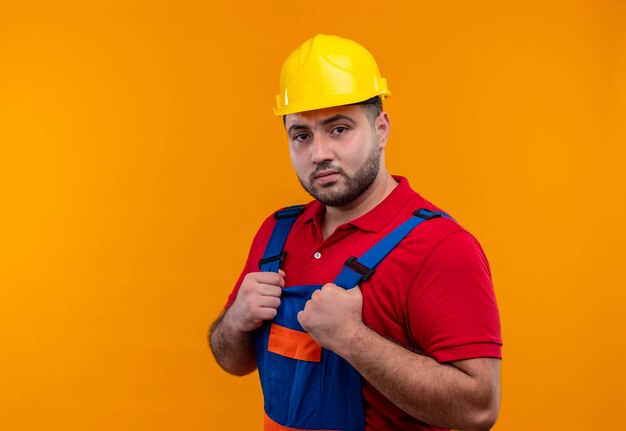 Young builder man in construction uniform and safety helmet holding hands on chest looking ar camera with serious confient expression 