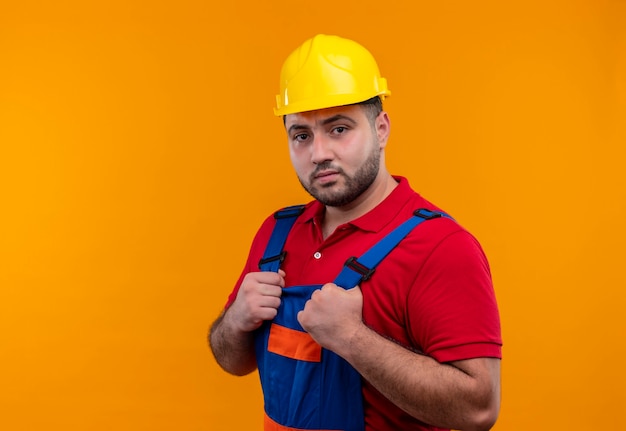 Молодой строитель в строительной форме и защитном шлеме, держась за руки на груди, глядя на камеру с серьезным выражением лица