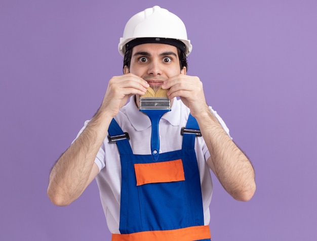 Giovane costruttore in uniforme da costruzione e casco di sicurezza che tiene il pennello imitando la barba divertendosi al lavoro in piedi sopra la parete viola