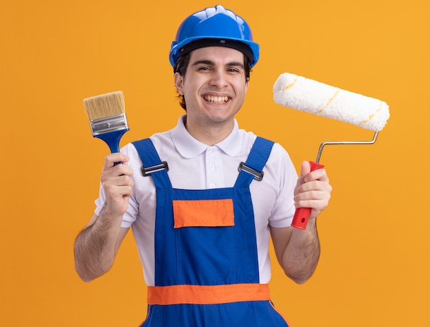 Молодой строитель в строительной форме и защитном шлеме, держа кисть и валик, глядя вперед, весело улыбаясь, стоя над оранжевой стеной