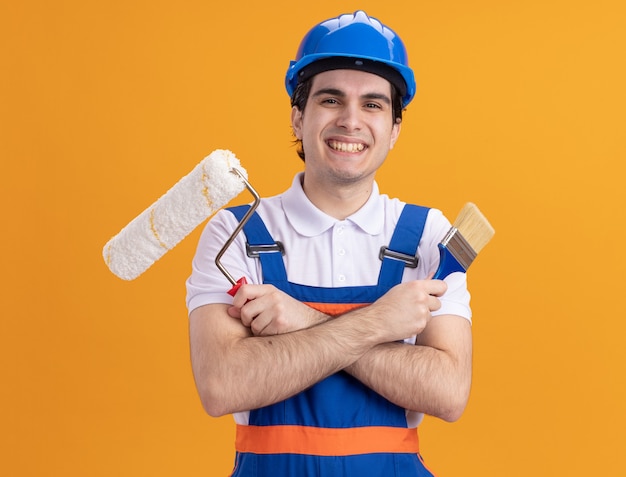 Young builder man in construction uniform and safety helmet holding paint brush and roller looking at front with big smile on face standing over orange wall