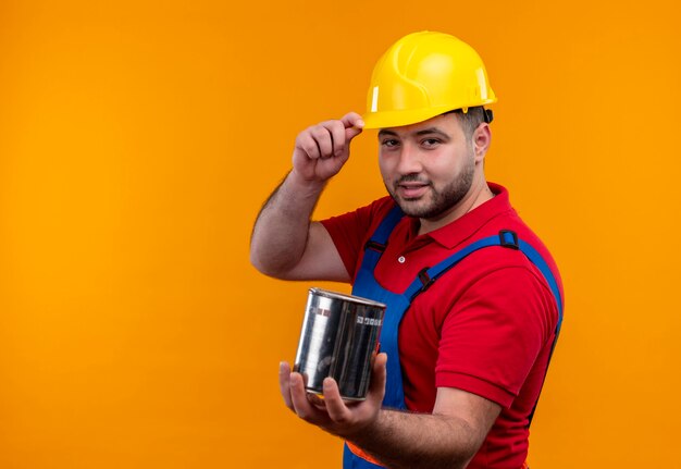 Young builder man in construction uniform and safety helmet holding paint can looking confident with smile touching helmet 